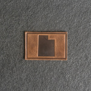 May include: A brown leather patch with the outline of the state of Utah in a darker brown color.