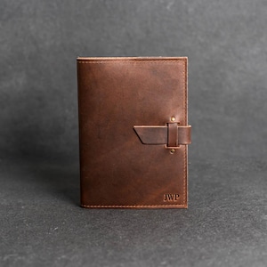 May include: A rectangular brown leather journal cover featuring a vertical strap closure secured with two brass rivets. The front cover displays the embossed initials JWP in the bottom right corner, with visible stitching along the edges.