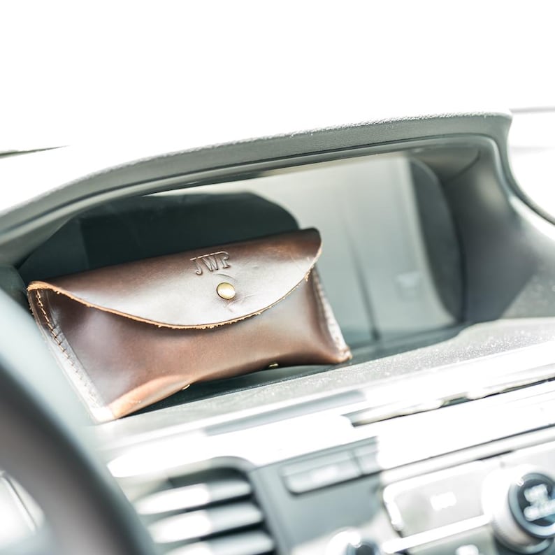 Puede incluir: Un estuche de cuero marr&oacute;n oscuro con cierre de bot&oacute;n a presi&oacute;n, grabado con las iniciales "JWP". El estuche rectangular con bordes redondeados est&aacute; en la consola de un coche. El cuero tiene un color rico y profundo.