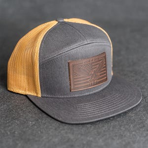 Könnte beinhalten: Graue und beige Trucker-Cap mit flachem Schirm. Die Kappe hat einen braunen Lederaufnäher mit einem amerikanischen Flaggen-Design und dem Text "1776-2024". Die Rückseite ist aus beigem Mesh.