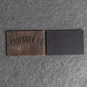May include: A brown leather patch with the text "PROPERTY OF John" in black lettering. The patch is attached to a black hook and loop fastener.
