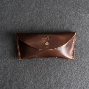Puede incluir: Un estuche de gafas de cuero marr&oacute;n oscuro con cierre de solapa y bot&oacute;n de lat&oacute;n. Las iniciales "JWP" est&aacute;n grabadas en la parte delantera. El estuche rectangular tiene bordes cosidos.