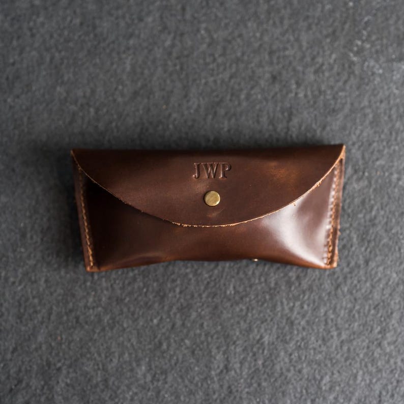 Puede incluir: Un estuche de gafas de cuero marr&oacute;n oscuro con cierre de solapa y bot&oacute;n de lat&oacute;n. Las iniciales "JWP" est&aacute;n grabadas en la parte delantera. El estuche rectangular tiene bordes cosidos.