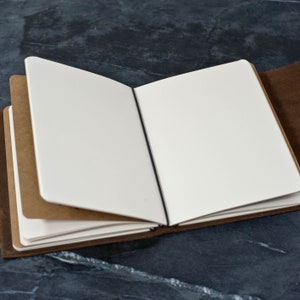 May include: A brown leather-bound journal with blank pages is open to reveal the white paper inside. The journal has a black ribbon bookmark.