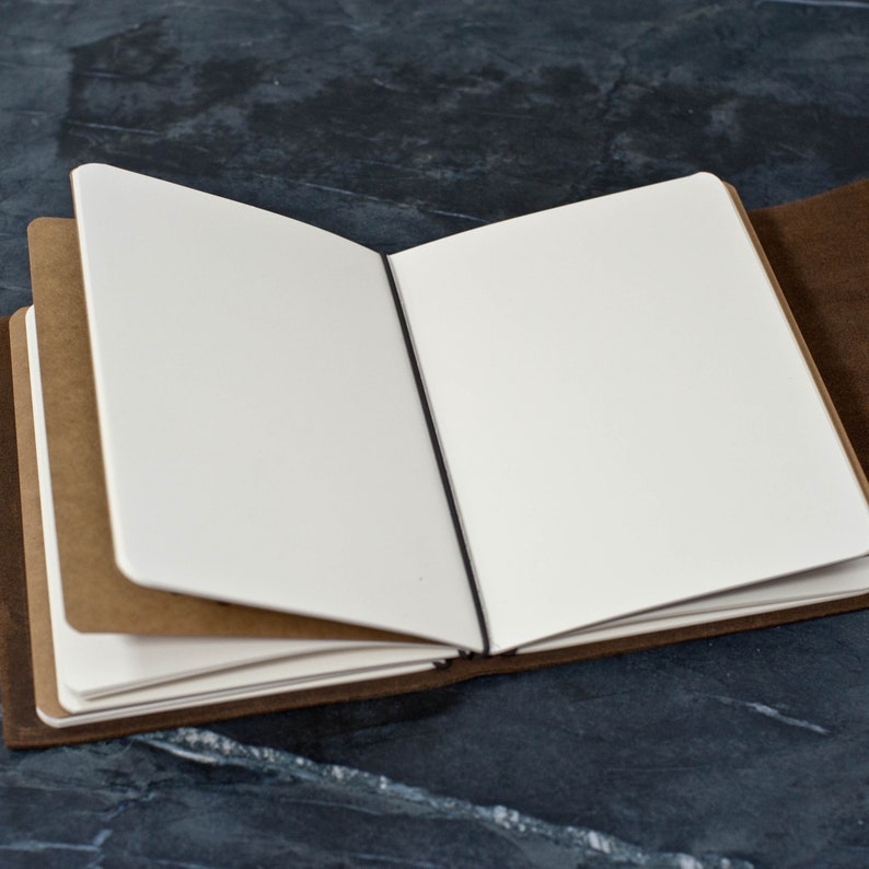 May include: A brown leather-bound journal with blank pages is open to reveal the white paper inside. The journal has a black ribbon bookmark.