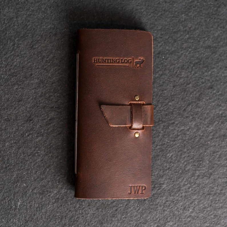 May include: A brown leather hunting log with the text "HUNTING LOG" and a deer graphic embossed on the cover. The log has a strap closure with brass rivets and the initials "JWP" at the bottom.