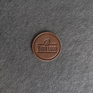 May include: A circular, brown leather patch featuring the text "YOUR LOGO" and a leaf motif. The patch is centrally positioned with a raised edge. The background is a dark grey, textured surface.