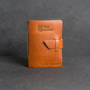 May include: A brown leather-bound journal with the text "5 Year Journal" embossed on the front cover. The journal has a leather strap closure and is personalized with the name "Taylor Anderson".