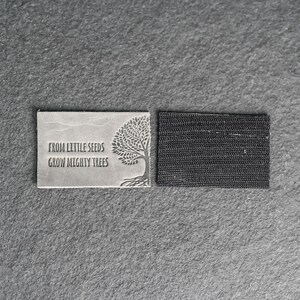 May include: A gray leather patch with the text "From little seeds grow mighty trees" and a stylized tree image. The patch has a black adhesive backing.