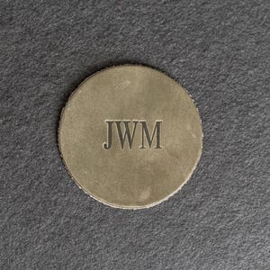 May include: A round, metallic, gold-colored disc with the initials "JWM" engraved in black lettering.