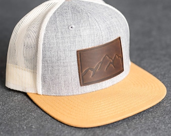 Puede incluir: Gorra de béisbol gris y amarilla con un parche de cuero marrón con un diseño de cordillera. La gorra tiene una parte trasera de malla blanca y una visera amarilla curvada. El botón superior también es amarillo.