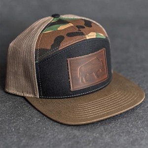 Op de afbeelding: Een pet met platte klep met een bruine klep en een zwart voorpaneel met een leren bisonpatch. De bovenkant van de pet heeft een camouflagepatroon en de zijkanten zijn van beige mesh.