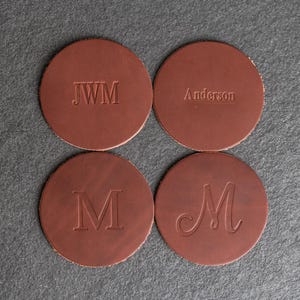 May include: Four brown leather coasters with different personalized engravings. The coasters feature the initials "JWM", the name "Anderson", and the letter "M" in two different styles.