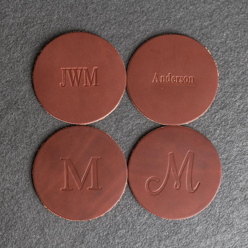 May include: Four brown leather coasters with different personalized engravings. The coasters feature the initials "JWM", the name "Anderson", and the letter "M" in two different styles.