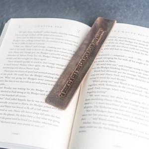 Peut inclure: Un marque-page en cuir marron avec le texte "A book is a gift you can open again and again" gravé dessus. Le marque-page est posé sur un livre ouvert.