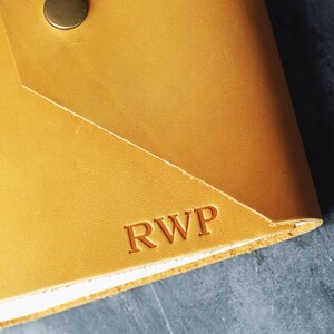 May include: A mustard yellow leather envelope-style wallet featuring a single metallic snap button closure at the top. The surface is embossed with the capital letters RWP in a serif font. The leather has a smooth texture and visible grain.