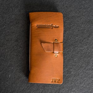 May include: A brown leather fishing log with the text "FISHING LOG" embossed at the top. The log has a strap closure with two brass rivets and the initials "JWP" at the bottom. The leather has a natural grain and a warm, inviting tone.