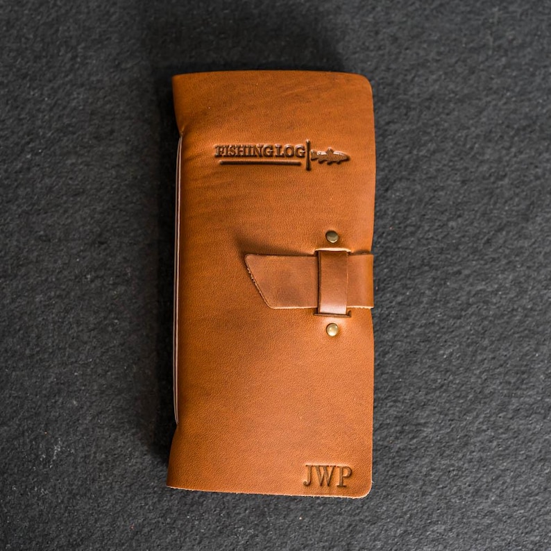 May include: A brown leather fishing log with the text "FISHING LOG" embossed at the top. The log has a strap closure with two brass rivets and the initials "JWP" at the bottom. The leather has a natural grain and a warm, inviting tone.
