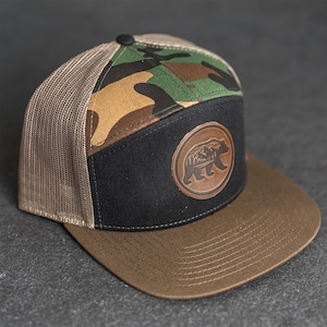 May include: A trucker cap with a brown peak and a black front panel featuring a circular leather patch with a bear and mountain design. The top has a camouflage pattern, and the mesh back is tan.