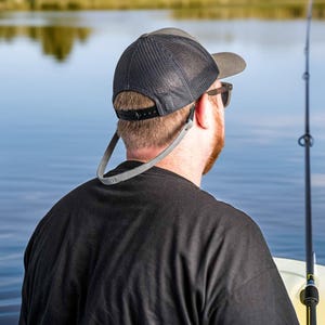 May include: A gray and black trucker hat with a mesh back and a gray strap. The hat is worn by a person wearing a black shirt, with a fishing rod in the background. The hat has a curved brim and a snapback closure.