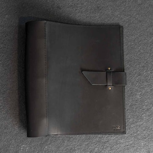 May include: A black leather portfolio with a flap closure and brass rivets. The portfolio has a stitched edge and the initials "TSR" are embossed on the lower right corner. The leather has a matte finish.