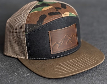 Puede incluir: Gorra de camionero con visera marrón y panel frontal negro con un parche de cuero marrón con un diseño de cordillera. La parte superior de la gorra tiene un estampado de camuflaje y la parte trasera es de malla.