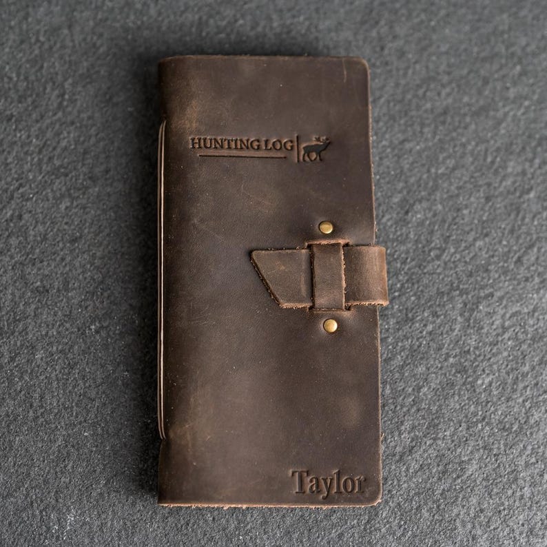 May include: A dark brown leather Hunting Log journal with the name "Taylor" embossed at the bottom. The journal has a strap closure with brass rivets and the words "HUNTING LOG" are embossed at the top.