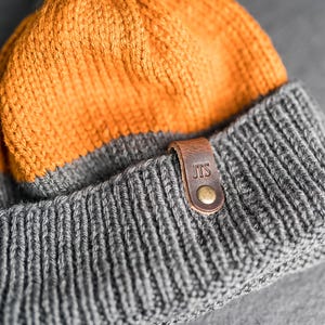 May include: A knit beanie featuring a two-tone design with a vibrant orange upper section and a dark gray ribbed cuff. A brown leather tag is attached to the cuff, embossed with the letters JTS and secured with a small metallic brass-toned rivet.