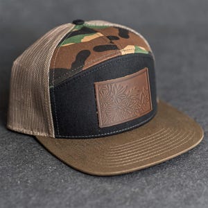 Op de afbeelding: Een bruine en zwarte baseballcap met een camouflagepatroon op de bovenkant. De cap heeft een bruin vizier, een zwart voorpaneel en een bruin leren patch met een bloemenmotief.
