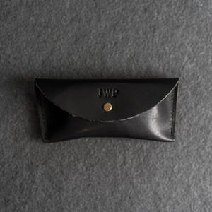 Puede incluir: Estuche de gafas de cuero negro con solapa curva y cierre de bot&oacute;n a presi&oacute;n dorado. Las iniciales "JWP" est&aacute;n grabadas en la parte delantera. El estuche es rectangular y parece hecho a mano.