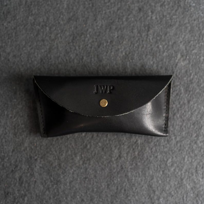 Puede incluir: Estuche de gafas de cuero negro con solapa curva y cierre de bot&oacute;n a presi&oacute;n dorado. Las iniciales "JWP" est&aacute;n grabadas en la parte delantera. El estuche es rectangular y parece hecho a mano.