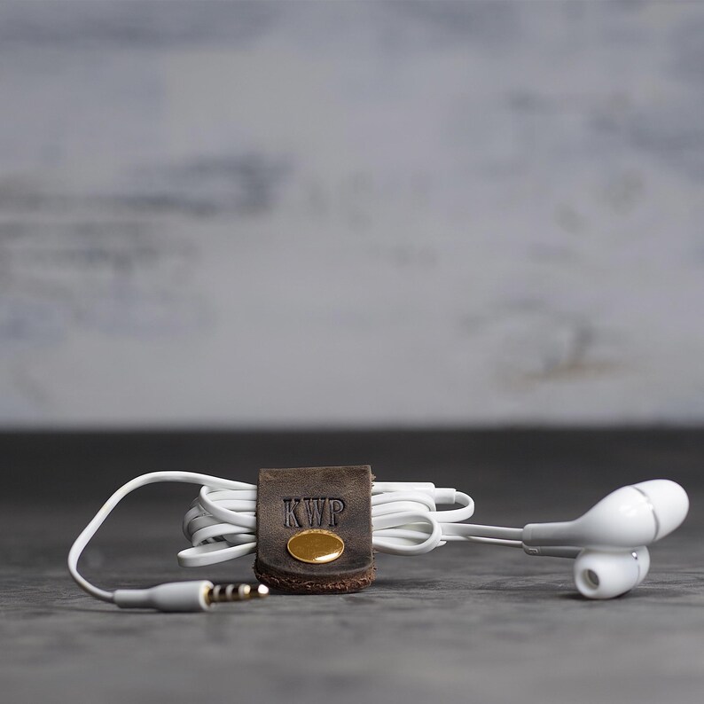 May include: A dark brown leather cord organizer with a gold-colored metal snap button and the letters KWP embossed on the front. The organizer is wrapped around a set of white wired earbuds with a standard 3.5mm audio jack connector.