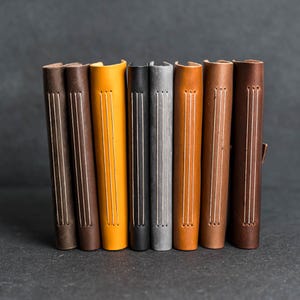 May include: A collection of handmade leather journals in various colors, including brown, yellow, black, and gray. Each journal features visible stitching along the spine and a textured leather finish. The journals are arranged vertically, showcasing their spines.