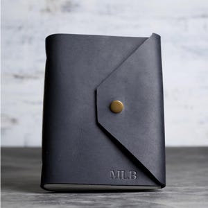 May include: A dark charcoal gray leather journal with an envelope-style flap closure secured by a single brass-colored metal snap button. The bottom right corner of the front cover features the embossed capital letters MLB in a serif font.