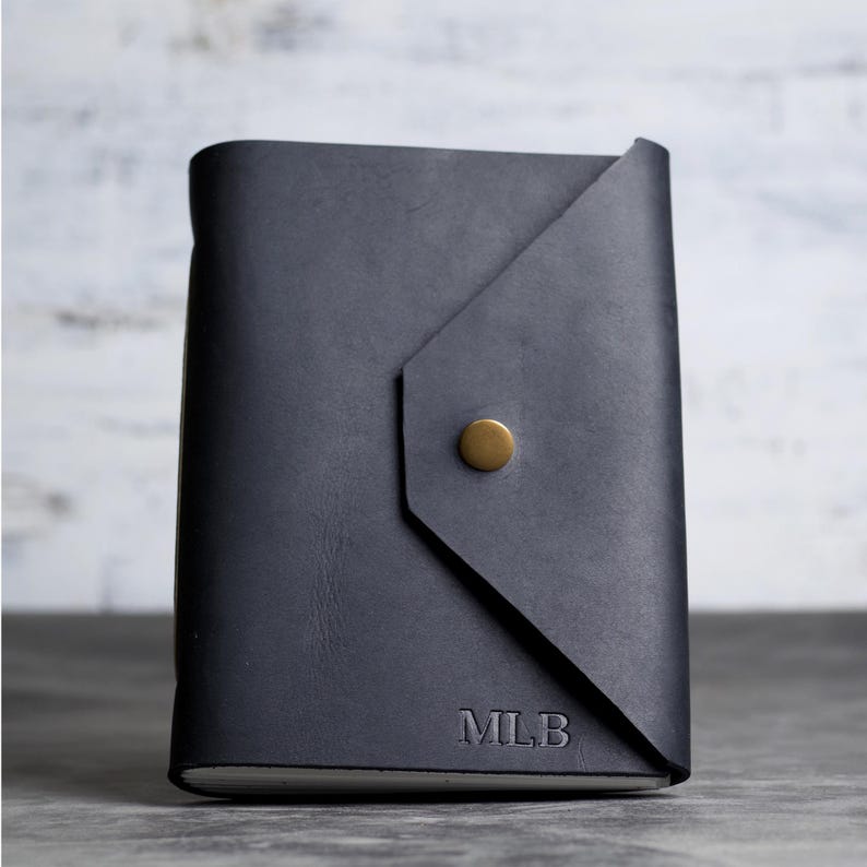 May include: A dark charcoal gray leather journal with an envelope-style flap closure secured by a single brass-colored metal snap button. The bottom right corner of the front cover features the embossed capital letters MLB in a serif font.