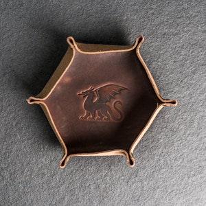 May include: A brown leather dice tray with a dragon design. The tray has six sides and is designed to hold dice.