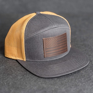 Peut inclure: Casquette de camionneur grise avec un écusson en cuir marron représentant le drapeau américain. La casquette a un dos en filet et une visière incurvée. Un accessoire élégant pour un look décontracté.