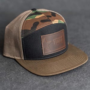 Op de afbeelding: Een bruine en zwarte truckerpet met een camouflage bovenkant en een bruine rand. De pet heeft een zwart voorpaneel met een bruin leren label met de tekst "TEXAS" met een ster. De pet heeft een mesh achterkant.