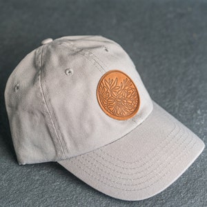 May include: A light gray baseball cap featuring a circular tan leather patch on the front panel. The patch is embossed with a detailed floral design consisting of various leaves and flower petals. The cap has a curved brim and stitched eyelets.