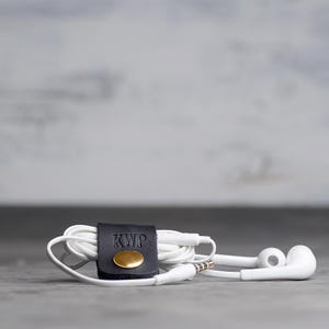 May include: A pair of white wired earbuds coiled and secured with a black leather cord organizer. The organizer features a gold-toned metal snap button and the embossed letters KWP on the front surface of the leather strap.