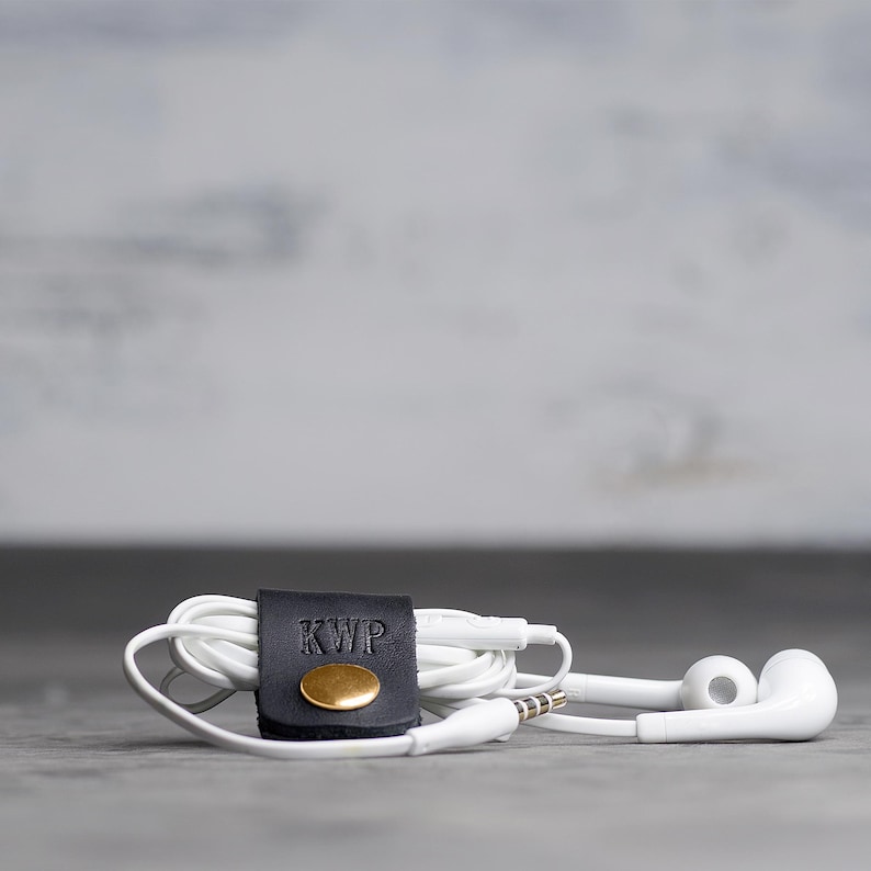 May include: A pair of white wired earbuds coiled and secured with a black leather cord organizer. The organizer features a gold-toned metal snap button and the embossed letters KWP on the front surface of the leather strap.