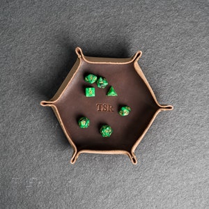 May include: A brown leather dice tray with a hexagonal shape and the word "TSR" embossed on the bottom. The tray is filled with green dice with gold numbers.