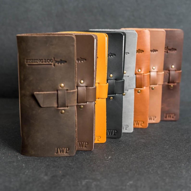 May include: A row of leather fishing logs in various colors, including brown, orange, and black. Each log has a strap closure and the text "FISHING LOG" with a fish graphic. The initials "JWP" or "WP" are embossed on the bottom.