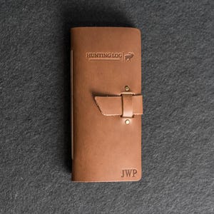 May include: A brown leather Hunting Log journal with a strap closure. The journal has the text "HUNTING LOG" and a deer graphic embossed on the cover. The initials "JWP" are embossed at the bottom.
