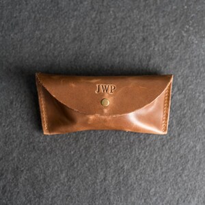 Puede incluir: Un estuche de gafas de cuero marr&oacute;n con las iniciales "JWP" grabadas en la solapa. El estuche es rectangular con una parte superior curvada y un cierre de bot&oacute;n a presi&oacute;n. Las costuras son visibles en los bordes.