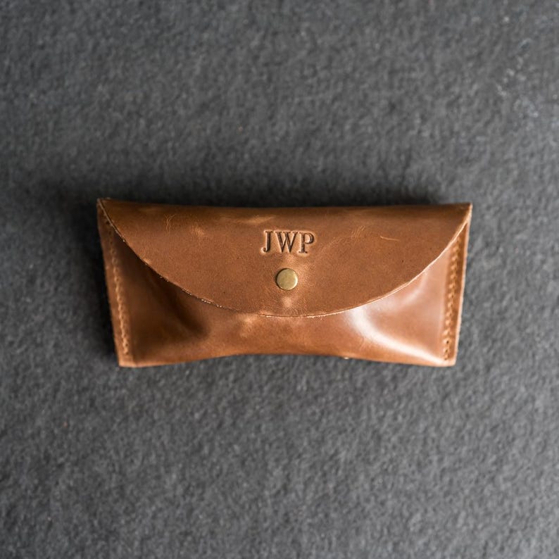 Puede incluir: Un estuche de gafas de cuero marr&oacute;n con las iniciales "JWP" grabadas en la solapa. El estuche es rectangular con una parte superior curvada y un cierre de bot&oacute;n a presi&oacute;n. Las costuras son visibles en los bordes.