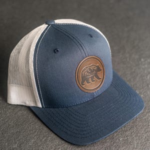 May include: A navy blue trucker hat with a white mesh back. The hat features a brown leather patch with a bear and mountain design. The hat has a curved brim and a button on top.