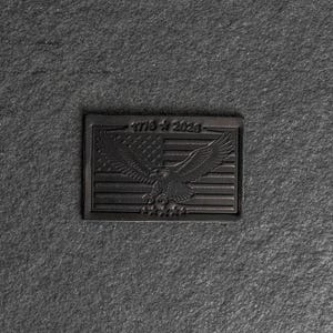 May include: A black rectangular emblem featuring an American flag design with an eagle and the text "1776-2023". The emblem is embossed with a detailed illustration of the flag and eagle.