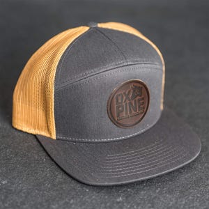 Può includere: Cappellino da camionista grigio e beige con visiera piatta. Il pannello frontale è grigio con una toppa rotonda in pelle con la scritta "OX & PINE LEATHER GOODS CO." Il retro in rete è beige.