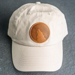 May include: A beige baseball cap featuring a circular brown leather patch on the front. The patch displays a debossed silhouette of a wolf howling at the moon. The cap has a structured crown, a curved brim with stitching, and a button on top.